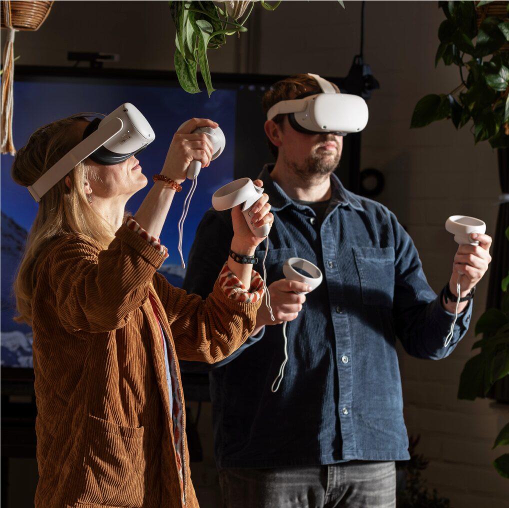 A woman in a brown jacket stands beside a man in a blue shirt. They both wear VR goggles.