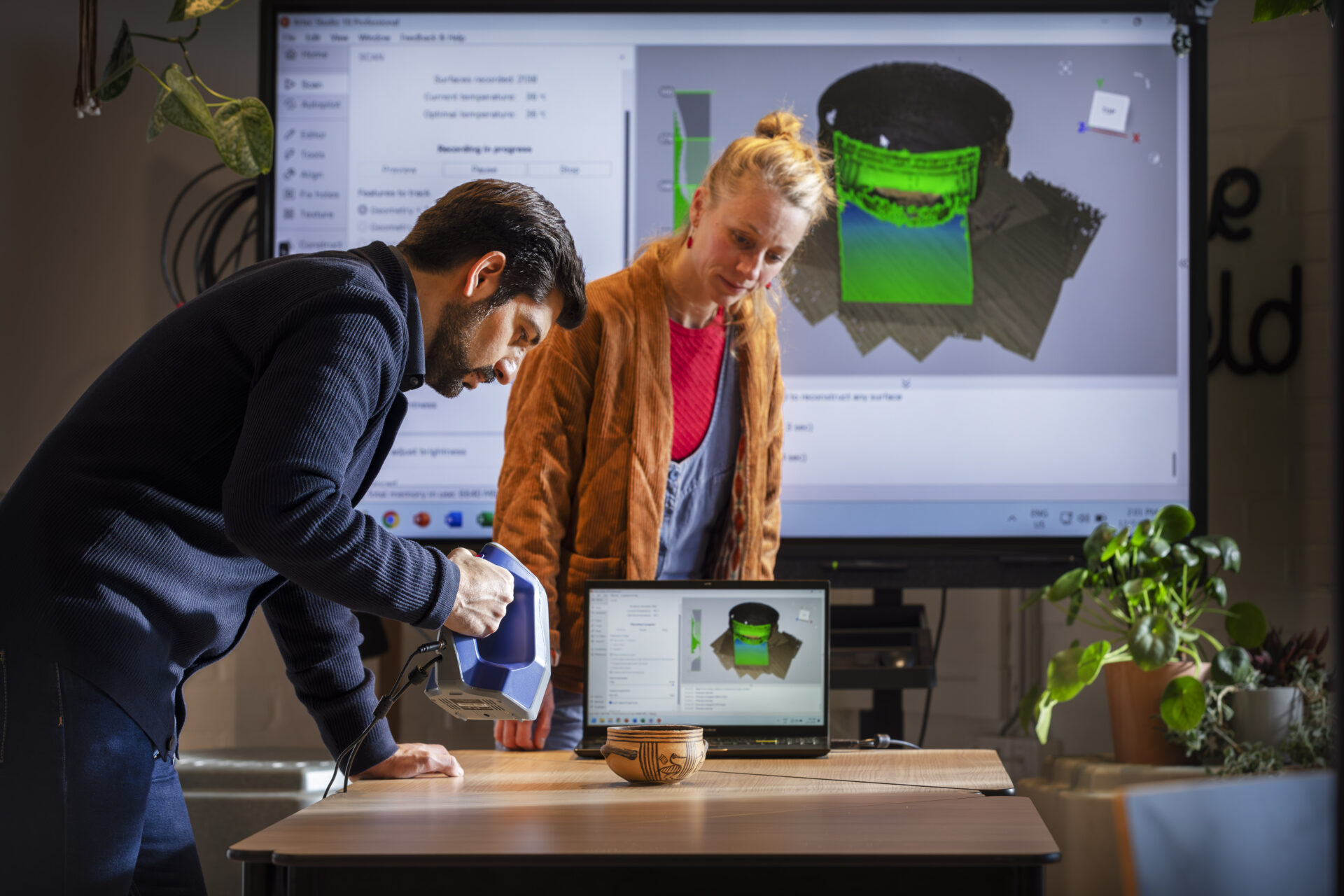 A man holds a 3D scanner in front of a screen. A blonde woman supervises.