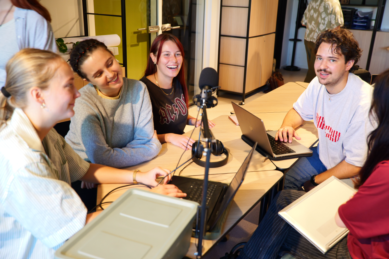 Photograph of 3 women and 1 man sitting around a table with a microphone in the middle, smiling and laughing looking at the camera